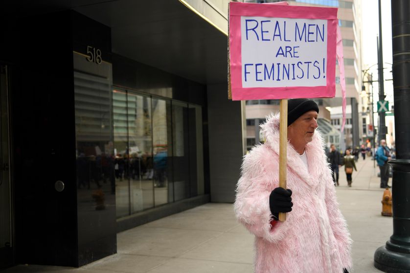Un homme dans la rue, habillé d'une sortie de bain rose bonbon, d'un bonnet et de gants, brandissant une pancarte en anglais proclamant : "Real Men Are Feminists"